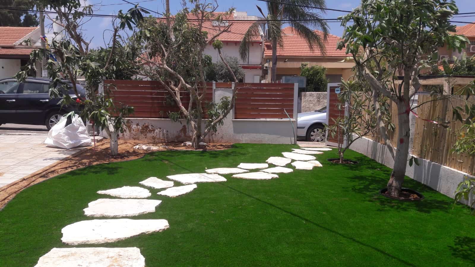 גינה קדמית עם דשא ירוק, עצים ושביל אבני דריכה לבנות.